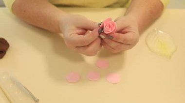 Masada oturan, yumuşak pembe polimer kil yapraklarıyla el yapımı gül yapan bir kadın.