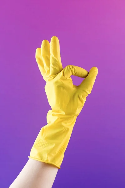Hand in yellow rubber glove shows OK gesture on purple background closeup. Professional cleaning service. Easy housework and routine chores