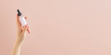 Açık pembe arka planda bir kadının elinde deri yağı olan cam bir damlacının maketi..