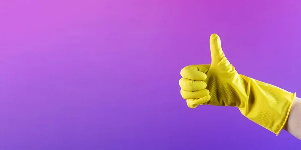 A hand in a yellow rubber glove for washing and cleaning shows a thumbs up on a purple background. Cleaning concept. Copy space