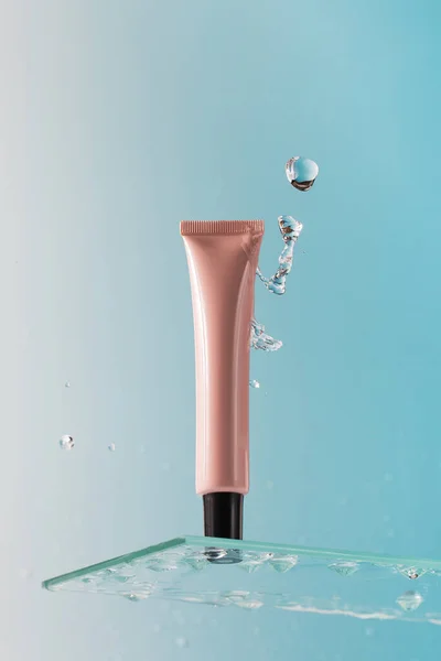 Mockup of a pink tube with cream on a glass shelf and splashes of water on a blue background close-up. Cosmetic product for mature skin. Place for a logo.