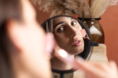 Aynanın yansımasındaki güzel esmer kız kuvars masaj rulosu kullanarak yüz masajı yapıyor. Evde cilt bakımı, yaşlanma önleyici cilt bakımı.