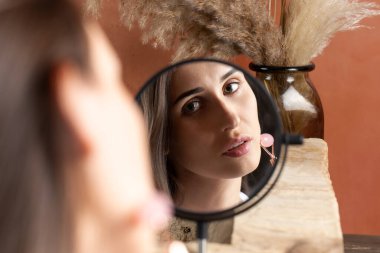 Aynanın yansımasındaki güzel esmer kız kuvars masaj rulosu kullanarak yüz masajı yapıyor. Evde cilt bakımı, yaşlanma önleyici cilt bakımı.