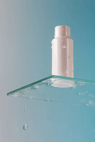 White tube of cosmetic product on a glass shelf and falling drops on a blue background. Skin care. Cosmetic product for body washing