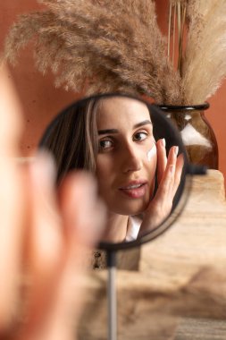 Aynanın önündeki genç bir kadın yüzüne pampa otlarının arka planına nemlendirici sürüyor. Güzellik portresi, günlük cilt bakımı kavramı.