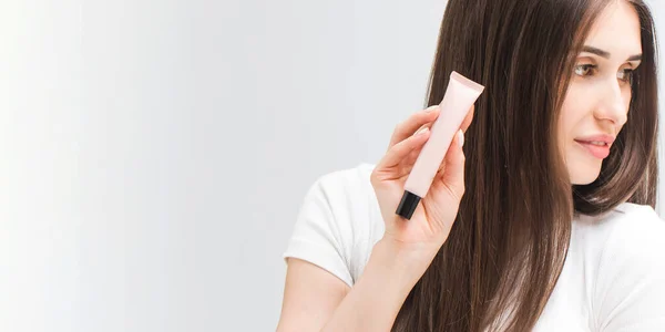 Woman with straight hair shows small tube of cosmetic product in studio closeup. Blank package of under-eye care product. Professional cosmetics. Banner