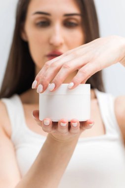 Genç bir esmer kadın, besleyici ve nemlendirici bir kremle büyük bir kavanoz açar. Stüdyoda hafif bir arka planda kozmetik ürünü. Organik bir ürün ve metin için bir yer