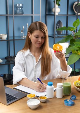 Genç diyet uzmanı turuncu yarısını tutuyor ve ofisteki dizüstü bilgisayarın yanında kağıda yazıyor. Doktor masasında farklı takviyeler var. Sağlıklı besin