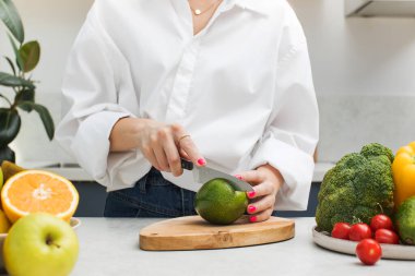 Genç bir kadın taze meyve ve sebzelerle çevrili mutfak masasında olgun bir avokadoyu ikiye böler. Vejetaryenlik kavramı, sağlıklı beslenme.