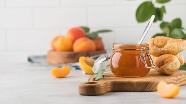 İçinde kaşık olan bir kavanoz kayısı reçeli, ahşap bir tabakta olgun kayısılar ve açık arkaplanda yeşil yapraklı bir dal. Kış için hazırlıklar, meyve koruma. Boşluğu kopyala