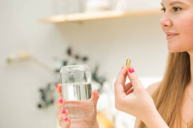 Mutfakta elinde bir bardak su ve bir kapsül balık yağı ya da D vitamini tutan mutlu sarışın kadın. Vitaminler ve besin takviyeleri. Sağlıklı bir yaşam tarzı. Koruyucu tıp.