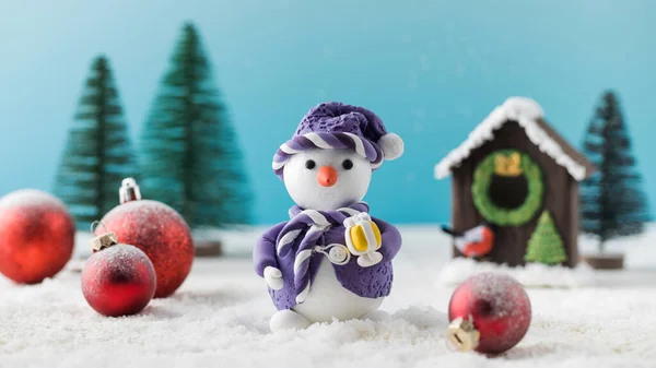 Funny toy snowman in purple clothes and red Christmas tree balls near small house in decorative forest on artificial snow close view
