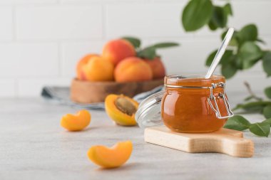 İçinde kaşık olan bir kavanoz kayısı reçeli, ahşap bir tabakta olgun kayısılar ve açık arkaplanda yeşil yapraklı bir dal. Kış için hazırlıklar, meyve koruma. Boşluğu kopyala