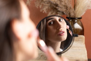 Aynanın yansımasındaki güzel esmer kız kuvars masaj rulosu kullanarak yüz masajı yapıyor. Evde cilt bakımı, yaşlanma önleyici cilt bakımı.