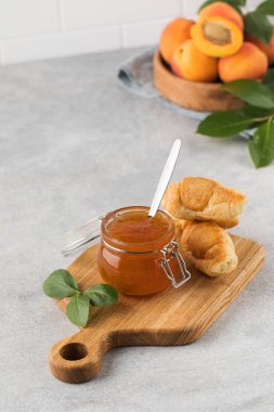 İçinde kaşık olan bir kavanoz kayısı reçeli, ahşap bir tabakta olgun kayısılar ve açık arkaplanda yeşil yapraklı bir dal. Kış için hazırlıklar, meyve koruma..