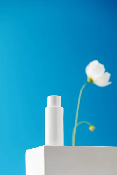 White tube with a cosmetic product on a podium on a blue background with a flower. The concept of natural plant cosmetics. Layout.