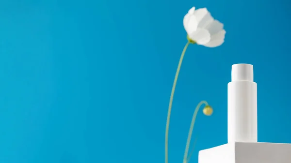 White tube with a cosmetic product on a podium on a blue background with a flower. The concept of natural plant cosmetics. Layout.