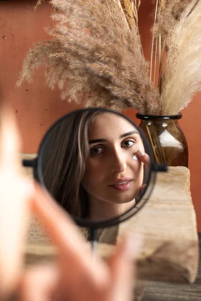 Aynanın önündeki genç bir kadın yüzüne pampa otlarının arka planına nemlendirici sürüyor. Güzellik portresi, günlük cilt bakımı kavramı.