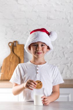 Noel Baba şapkalı ve beyaz tişörtlü şirin küçük çocuk ev yapımı kurabiyeleri açık mutfak manzaralı süt bardağına koyar.