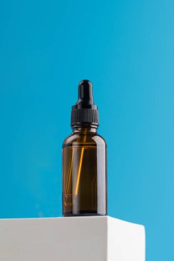 Mavi arka planda bir podyumda kozmetik ürünü olan kahverengi bir cam damlası. Cilt bakımı.