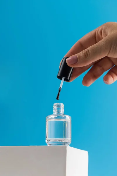A womans hand takes out a brush from a jar of transparent medical nail polish. Transparent nail polish stands on a podium on a blue background.