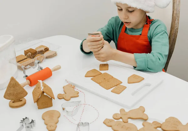 かわいい男の子が大晦日のサンタの帽子のテーブルでジンジャーブレッドクッキーを飾ります 子供とクリスマス休暇 — ストック写真