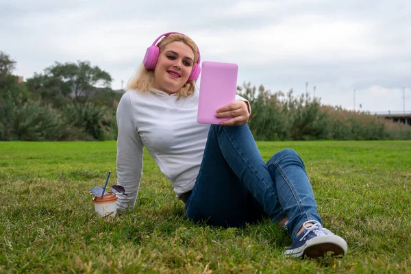 Parktaki çimenlerde oturan genç bir kadın pembe kulaklıklı pembe bir tablete bakıyor..
