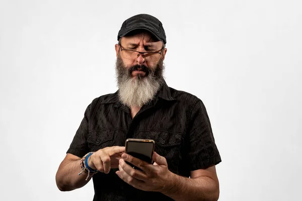 Vuxen man med vitt skägg, casual kläder och glasögon kisar för att se mobilen. — Stockfoto