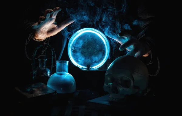 Gros plan des mains d'un magicien racontant l'avenir avec une boule de cristal bleue, entourée de fumée, de livres, d'un crâne et d'éléments mystiques. Photos De Stock Libres De Droits