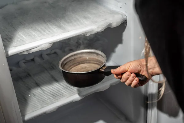 냉장고에 있는 얼음을 식히기 위해 뜨거운 물을 담은 냄비를 손으로 들고 스톡 이미지