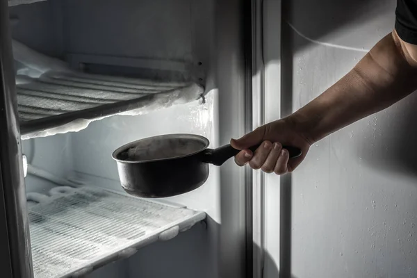 Main introduisant une casserole avec de l'eau chaude pour dégeler la glace dans le réfrigérateur. Image En Vente
