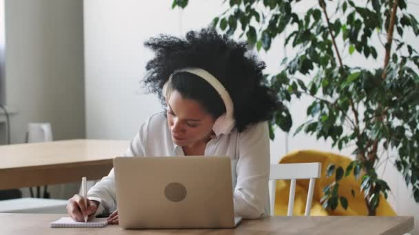 非洲裔美国妇女在电话会议上使用笔记本电脑和耳机在记事本上记笔记的肖像。布鲁内特坐在总公司的桌旁。靠近点慢动作准备59.97fps. — 图库视频影像