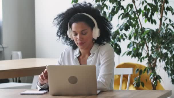 Portret Afroamerykanki rozmawiającej przez wideo-konferencję przy użyciu laptopa i słuchawek robiącej notatki na notatniku. Brunetka siedzi przy stole w biurze. Zamknij drzwi. Wolny ruch gotowy 59.97fps. — Wideo stockowe