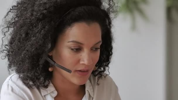 Portrait d'une jeune femme afro-américaine parlant par vidéoconférence à l'aide d'un casque. Brunette aux cheveux bouclés en chemisier blanc s'assoit à table au bureau à la maison. Ferme là. Ralenti prêt 59.97fps. — Video