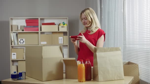 Trabajador de almacén está trabajando en una sala de oficina contra el fondo de las cajas. Mujer prepara paquetes para la entrega, mensajes de texto en el teléfono inteligente, se regocija por las buenas noticias. De cerca. Cámara lenta lista 59.94fps. — Vídeos de Stock