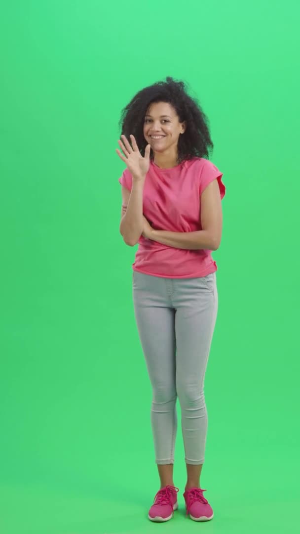 Портрет молодой женщины афроамериканки кокетливо улыбающейся, машущей рукой и показывающей жест сюда. Черная женщина с вьющимися волосами позирует на зеленом экране. Slow motion ready 59.97fps. Полная длина. — стоковое видео