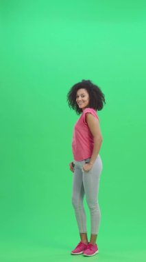 Genç Afro-Amerikan kadın portresi kameraya flört ederek gülümsüyor ve göz kırpıyor. Kıvırcık saçlı siyah kadın yeşil ekranda poz veriyor. Yavaş çekim hazır 59.97 fp. Dikey video. Tam uzunluk.