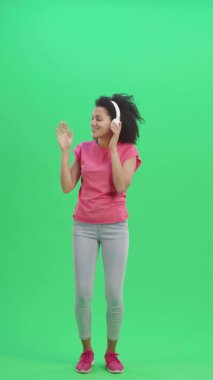 Genç Afro-Amerikan kadın dansının portresi ve büyük beyaz kulaklıklarla müzik dinlemenin keyfi. Kıvırcık saçlı siyah kadın yeşil ekranda poz veriyor. Yavaş çekim hazır 59.97 fp. Dikey video. Tam uzunluk