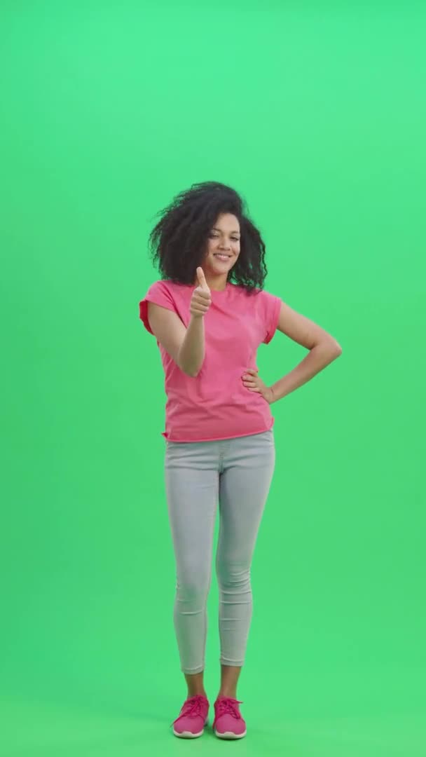Porträtt av unga kvinnliga afroamerikaner som visar tummen upp, gester som. Svart kvinna med lockigt hår poserar på grön skärm i studion. Slow motion klar 59,97fps. fp. Vertikal video. Full längd. — Stockvideo