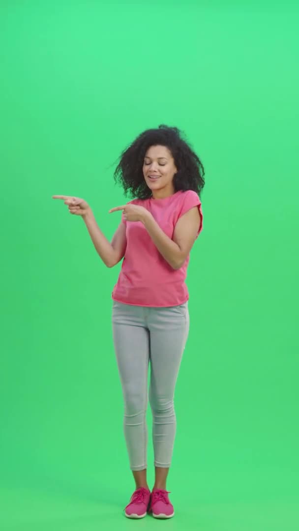 若い女性のアフリカ系アメリカ人の肖像画は、次に何かを指摘し、親指のようなものを示しています。黒人女性は緑色の画面にポーズをとる。スローモーション準備ができて59.97fps。垂直ビデオ。全長. — ストック動画