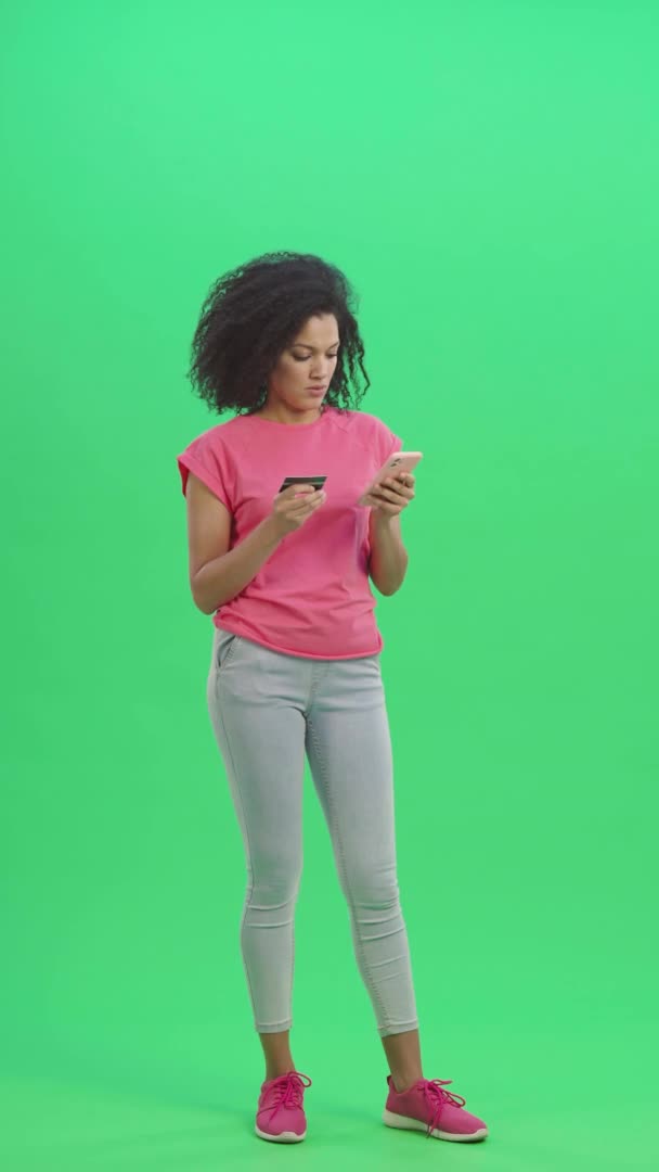 Portrait of young female African American makes an online purchase using credit card and phone. Black woman poses on green screen. Slow motion ready 59.97fps. Vertical video. Full length. — Stock Video