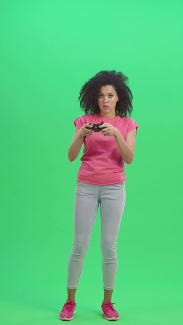 Porträtt av unga kvinnliga afroamerikaner som spelar ett tv-spel med hjälp av en trådlös styrenhet. Svart kvinna med lockigt hår poserar på grön skärm. Slow motion klar 59,97fps. fp. Vertikal video. Full längd. — Stockvideo
