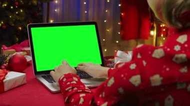 Kadın evde Noel ağacının önünde krom renkli yeşil ekranlı dizüstü bilgisayarla alışveriş yapıyor. Dişi, internetten noel hediyesi alıyor. Kutlama konsepti. Kapatın. Yavaş çekim.