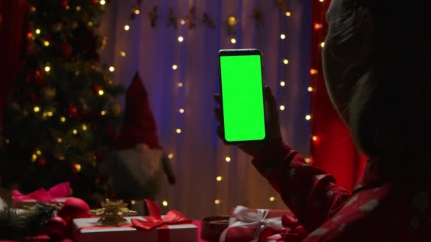 여성이 새 년 이브 (New Years Eve) 에 장식 된 집 내부 배경에 녹색 스크린 크롬 키가 달린 스마트폰을 손에 들고 있다. 비디오 채팅을 통한 온라인 커뮤니케이션. 닫아. 느린 동작. — 비디오