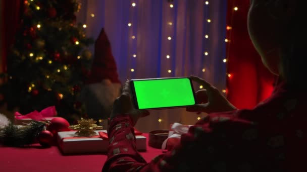 여성은 크리스마스 장식, 조명, 새해 트리등 배경을 놓고 앉아 녹색 스크린 크로마 키를 가진 스마트폰상의 콘텐츠를 보고 있다. 닫아. 느린 동작. — 비디오
