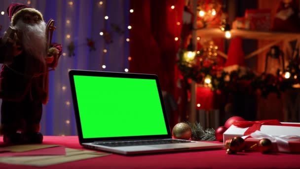 Laptop z zielonym ekranem chroma klucz stoi na czerwonym stole obok Świętego Mikołaja, prezenty i zabawki. Dom pokój z wystrojem świątecznym i światła w rozmytym tle. Monitor makiety. Zamknij drzwi. Zwolniony ruch. — Wideo stockowe
