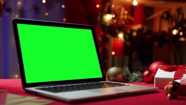Yeşil ekran krom anahtarı olan bir dizüstü bilgisayar hediye ve oyuncakların yanında kırmızı bir masada duruyor. Noel dekorlu, arka planda bulanık ışıklar olan ev odası. Reklam için yer var. Kapatın. Yavaş çekim — Stok video