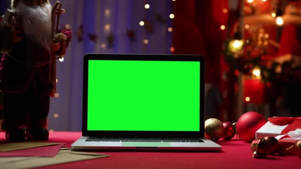 Laptop z zielonym ekranem chroma klucz stoi na czerwonym stole obok Świętego Mikołaja, prezenty i zabawki. Dom pokój z wystrojem świątecznym i światła w rozmytym tle. Monitor makiety. Zamknij drzwi. Zwolniony ruch. — Wideo stockowe