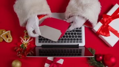 Noel Baba 'nın elleri beyaz eldivenlerle kırmızı yeni yıl süsleme masasında klavye bilgisayarında daktilo ediyor. Noel Baba çocuklardan gelen mektuplara bakar ve internetten hediyeler alır. Kapatın. Yavaş çekim.