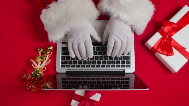 Top view Santa ręce w białych rękawiczkach wpisując na klawiaturze przez czerwony Nowy Rok urządzony stół. Święty Mikołaj pracuje z laptopem, przegląda pocztę i odpowiada na wiadomości do dzieci. Zamknij drzwi. Zwolniony ruch. — Wideo stockowe